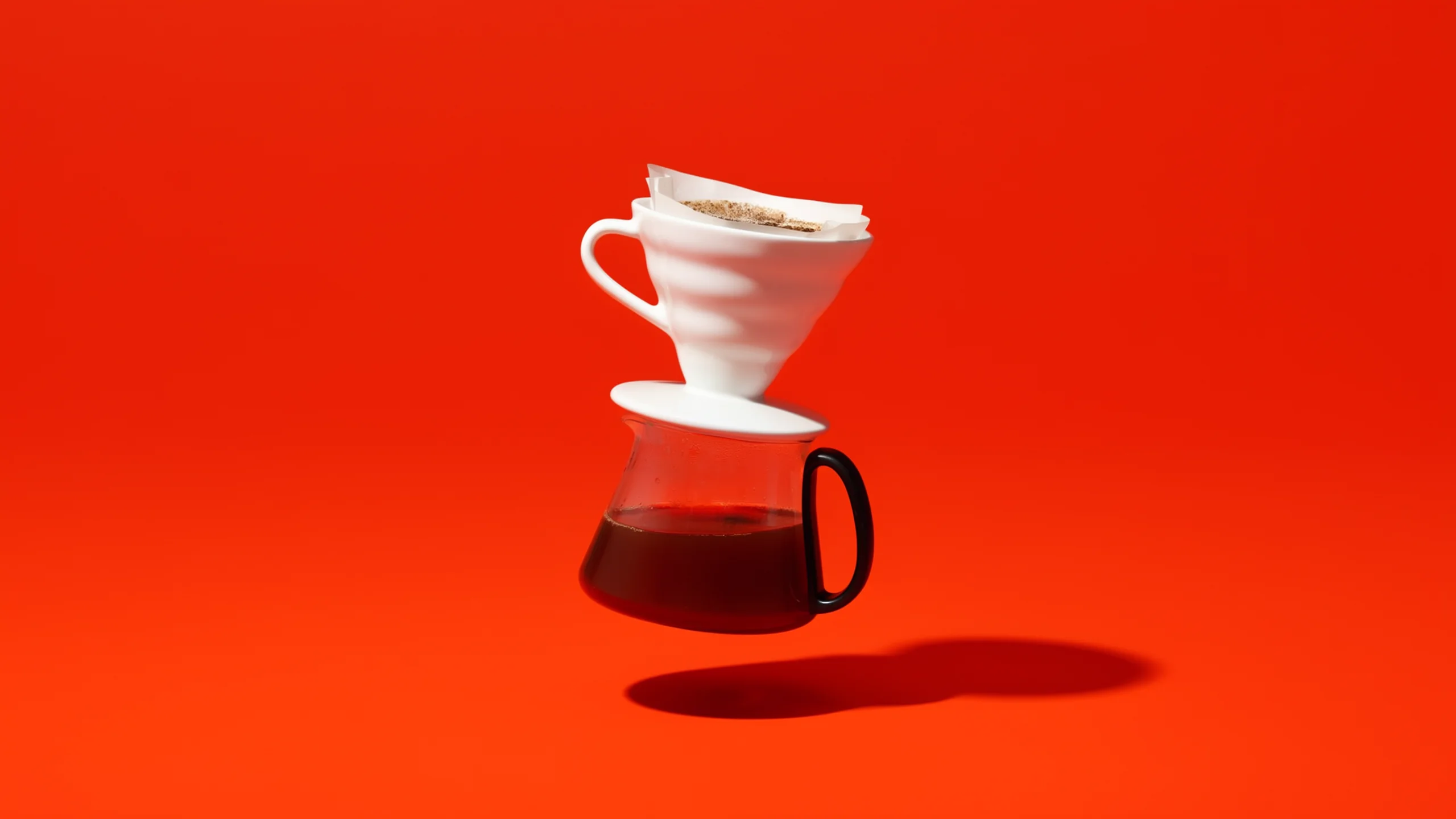 White ceramic pour-over dripper and glass coffee server floating against a vivid orange studio background.